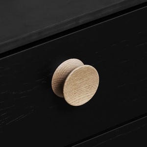 May include: A close-up shot of a black wooden chest of drawers featuring two circular, natural wood knobs. The knobs are light-coloured, providing a contrast against the dark wood. The wood grain is visible.