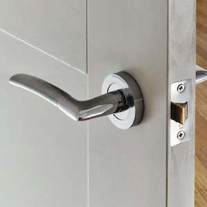May include: A polished chrome door handle with a curved lever design. The handle is mounted on a light grey door, with a matching chrome door latch visible. The door appears to be made of wood.