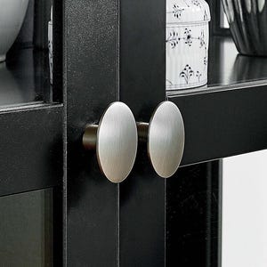 May include: Two silver-toned cabinet door knobs with a brushed finish are mounted on a black cabinet door.