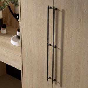 May include: Close-up of a light wood grain cabinet door with two long, black metal handles. The handles are vertical and have a minimalist design. The cabinet door is part of a larger furniture piece, possibly a wardrobe or dresser.
