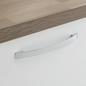 May include: A polished chrome cabinet pull handle with a curved design. It's fixed to a white cabinet door, with a light brown wooden countertop above. The handle is approximately 15 cm in length.