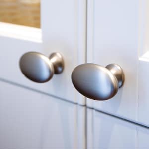 May include: Two brushed nickel oval cabinet knobs on a white cabinet door. The knobs have a smooth, rounded shape and a metallic finish, offering a modern look. The cabinet door is clean and simple.