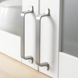 May include: Two brushed nickel cabinet pulls with a rectangular shape and rounded edges. The pulls are mounted on a white cabinet door.