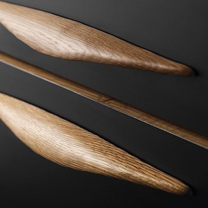 May include: Two wooden drawer pulls with a curved, elongated shape. The pulls are made of light brown wood with a natural grain pattern. The pulls are mounted on a black surface.