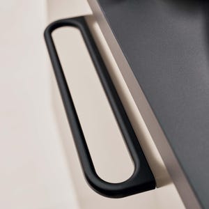 May include: Close-up of a black, U-shaped handle attached to a dark grey surface. The handle has a smooth, matte finish and is mounted on a light beige wall. The design is modern and minimalist.