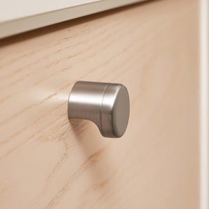 May include: A close-up shot of a brushed nickel cabinet knob. The knob has a cylindrical body with a rounded face and a curved base, attached to a light wood grain cabinet door. The hardware offers a modern look.