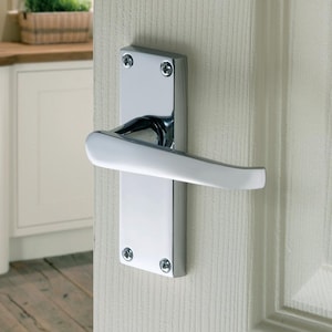 May include: A shiny chrome door handle, featuring a rectangular backplate and a curved lever. The handle is fixed to a white door with a textured surface. The backplate is secured with four screws.