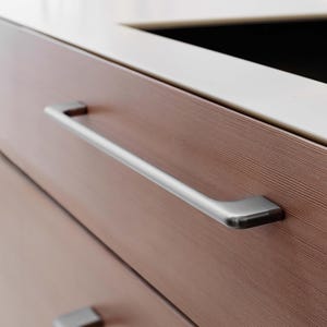 May include: Close-up of a brown wooden cabinet drawer with a long, brushed silver handle. The drawer has a wood grain texture. A white countertop is visible in the background.