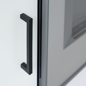 May include: A black metal door handle with a rectangular shape. The handle is attached to a white door.