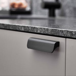 May include: A close-up of a kitchen cabinet. The light grey cabinet door features a dark grey, curved handle. The countertop is speckled grey granite. The background is blurred, showing a kitchen setting.