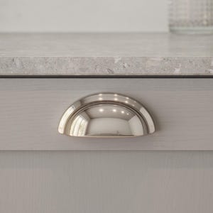 May include: A polished nickel cabinet pull in a half-moon shape. The handle is mounted on a light grey cabinet door. The reflective surface of the pull mirrors the surroundings. A classic design for kitchen or bathroom cabinets.