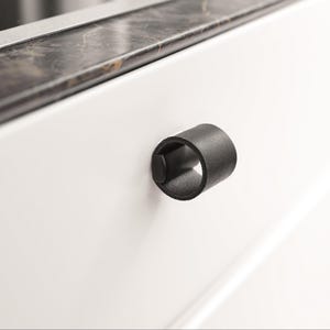 May include: A black leather drawer pull with a loop design, mounted on a white cabinet door. The cylindrical pull is shown in close-up. The cabinet has a marble countertop.