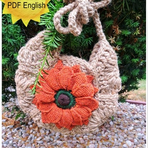May include: A crocheted bag made from natural jute twine with a large orange flower appliqué. The bag has a long strap and is hanging from a tree branch.