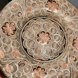 May include: Ornate, decorative plate with intricate floral and scrollwork patterns. The plate features a central design and is embellished with copper-colored accents. The overall color scheme is a mix of silver and copper tones, creating a detailed and elegant appearance.