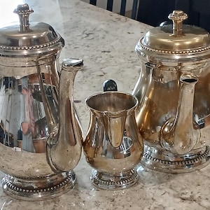 Könnte beinhalten: Ein silbernes Teeservice mit Kaffeekanne, Teekanne und Sahnekännchen. Die Kaffeekanne und die Teekanne haben schwarze Griffe und dekorative Perlenverzierungen. Das Sahnekännchen ist kleiner und schlicht gestaltet.