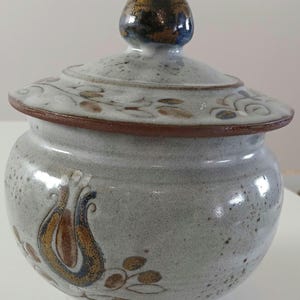 May include: A white ceramic jar with a lid, decorated with brown, gold, and blue floral designs. The lid has a knob in the shape of a rounded, brown and gold bulb.