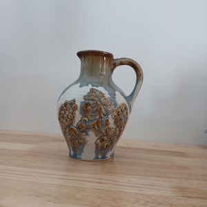 Pode incluir: Um jarro de cerâmica com corpo bulboso e uma única alça. A superfície é esmaltada em branco com detalhes em azul e castanho. O exterior é decorado com padrões em relevo moldados de cachos de uvas e folhas de videira.