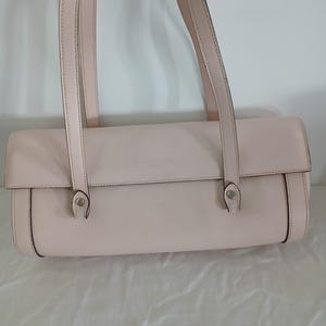 May include: A pale pink leather handbag with two long straps. The rectangular bag has rounded edges and a flap closure. The straps are attached with small metal accents. The bag is displayed against a white background.