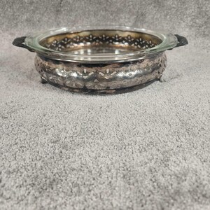 May include: A silver serving dish with a clear glass insert. The dish has a decorative, hammered metal exterior with a scalloped edge and small feet. Black handles are on either side of the glass insert.