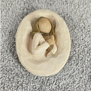 Könnte beinhalten: Geschnitzte Holzfigur einer Person, die ein kleines Tier hält, in einem ovalen Rahmen. Die Figur hat blondes Haar und trägt ein weißes Kleidungsstück. Das Tier ist braun. Der Rahmen hat ein strukturiertes, geschnitztes Aussehen.