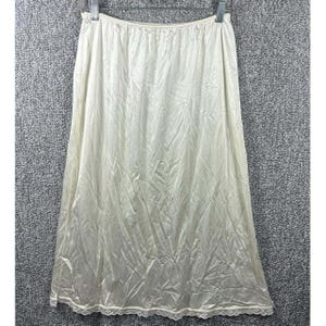 May include: Ivory-colored half slip with a satin finish, hanging from a metal hanger. The slip has a straight cut, reaching mid-calf length, and features a delicate lace trim along the hemline. The fabric has subtle vertical lines.