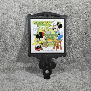 May include: A black metal trivet with a square ceramic tile featuring Mickey and Minnie Mouse in a kitchen scene. Mickey wears a chef's hat and Minnie holds a pie. The tile is set in a decorative frame.