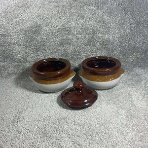 May include: Two small ceramic pots with lids, featuring a two-tone design with a dark brown glazed rim and upper section, transitioning to a light beige or off-white base. Each pot has small side handles and one matching dark brown lid.