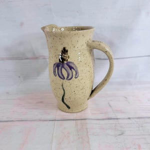 Può includere: Brocca in ceramica beige macchiata con manico e beccuccio. La brocca presenta un motivo floreale viola e verde dipinto a mano sul davanti. L'articolo è fatto a mano.