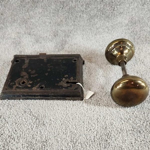 May include: A rectangular black metal door lock plate with visible wear and screw holes, positioned next to a brass door knob set featuring two round knobs connected by a central metal spindle, showing a metallic finish and a classic hardware design.