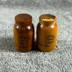 Może przedstawiać: Dwie drewniane solniczki i pieprzniczki w kształcie miniaturowych słoików typu Mason. Każdy pojemnik ma ciemnobrązowe wykończenie i zakręcaną pokrywkę z otworami. Z przodu widnieje nadruk Mason Salt Jar lub Mason Pepper Jar oraz Forge, Pa.