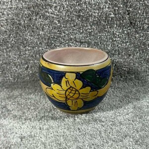 May include: A small, round ceramic bowl with a yellow and blue floral design. The bowl features a yellow flower with a blue background and a yellow rim. The interior is a light pink color.