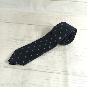 May include: A navy blue tie with a repeating pattern of small, light blue floral designs. The tie is made of a smooth fabric and is lying on a light wood surface.