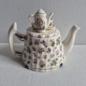 Puede incluir: Una tetera de cerámica color crema con un delicado patrón floral de flores rosas y verdes. La tapa está rematada con una figura en miniatura de una tetera y una taza. La tetera tiene un cuerpo acanalado, un asa curva y un pico vertedor.