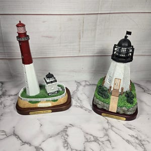 Op de afbeelding: Twee decoratieve vuurtorenbeeldjes. Eén is rood en wit, met een kleine aangrenzende structuur, en de andere is wit met een zwart dak. Beide staan op houten sokkels met identificatietekst.