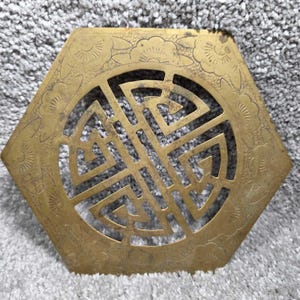 May include: Hexagonal brass trivet with a central geometric design and floral border. The trivet has a cut-out pattern and a warm, aged appearance. The design is likely of Asian origin.