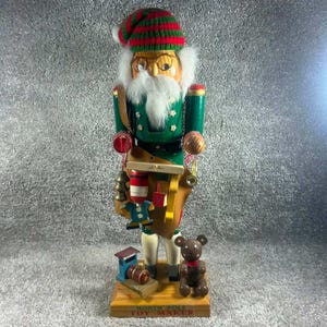 Op de afbeelding: Een houten notenkraker speelgoedmaker figuur met een groene jas en een rood-groen gestreepte muts. Het figuur heeft een witte baard, een bril en houdt miniatuurspeelgoed vast. Een kleine blauwe trein en een bruine teddybeer staan op de voet met NORTH POLE TOY MAKER.