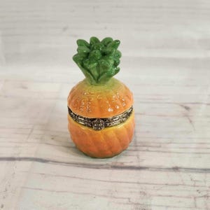 May include: A decorative pineapple-shaped trinket box. The box is orange with a green leafy top. The hinged lid is accented with silver-colored metal. The box is sitting on a white and gray wooden surface.