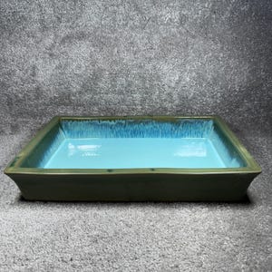 Puede incluir: Una jardinera rectangular de cerámica con interior azul claro y exterior verde oscuro. La jardinera tiene un acabado brillante y una superficie ligeramente texturizada. Los bordes de la jardinera son redondeados y el diseño general es simple y moderno.