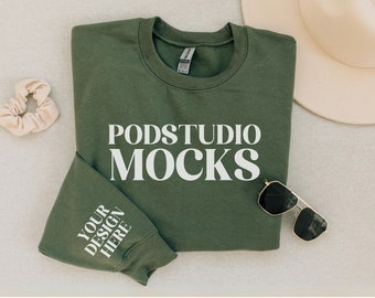 May include: A green sweatshirt with the text "PODSTUDIO MOCKS" printed on the front and "YOUR DESIGN HERE" printed on the sleeve. The sweatshirt is laid flat on a light beige surface with a hat and sunglasses.