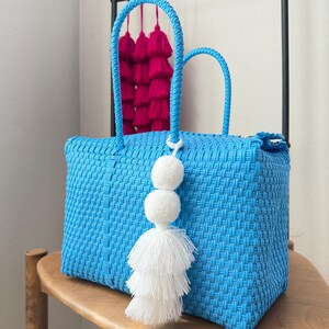 May include: A bright blue woven tote bag with matching handles. The bag is decorated with a white tassel and pom-pom. In the background, a rack displays pink tassels. The bag sits on a wooden chair.