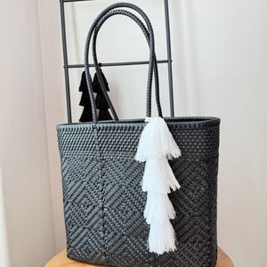 May include: A black woven tote bag featuring a rectangular shape with a chevron pattern throughout. The bag has two braided handles and is decorated with a large, multi-layered white tassel attached to one of the handles near the top opening.