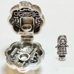 Puede incluir: Un relicario plateado abierto, que revela un anillo y herramientas en miniatura en su interior. Junto a él, una pequeña figura plateada de una persona con una capa con capucha. El relicario tiene forma de flor.
