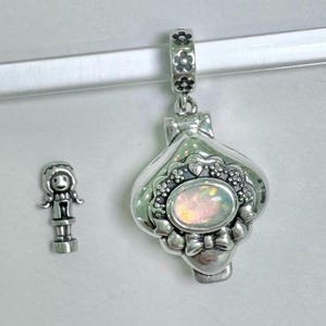 May include: A silver charm with a heart-shaped locket that opens. The locket has a white opal-like stone in the center and is decorated with floral and heart designs. A small silver charm of a girl with a dress and pigtails is also shown.