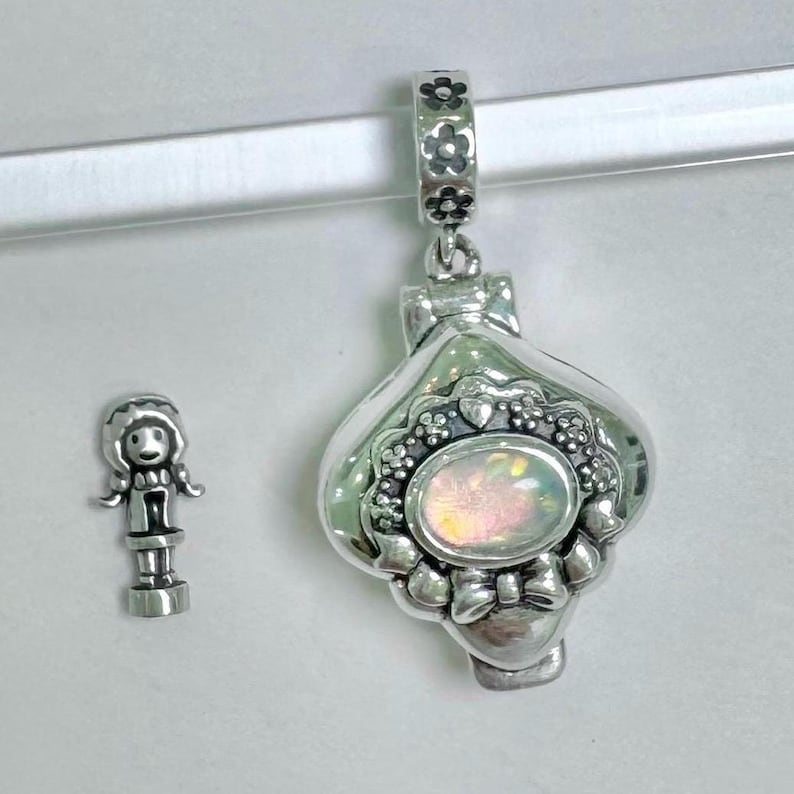 May include: A silver charm with a heart-shaped locket that opens. The locket has a white opal-like stone in the center and is decorated with floral and heart designs. A small silver charm of a girl with a dress and pigtails is also shown.