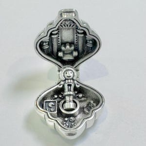 Pu&ograve; includere: Un ciondolo d'argento a forma di casetta delle bambole con una piccola figura di bambola all'interno. La casetta delle bambole &egrave; aperta e l'interno &egrave; decorato con mobili e giocattoli in miniatura.