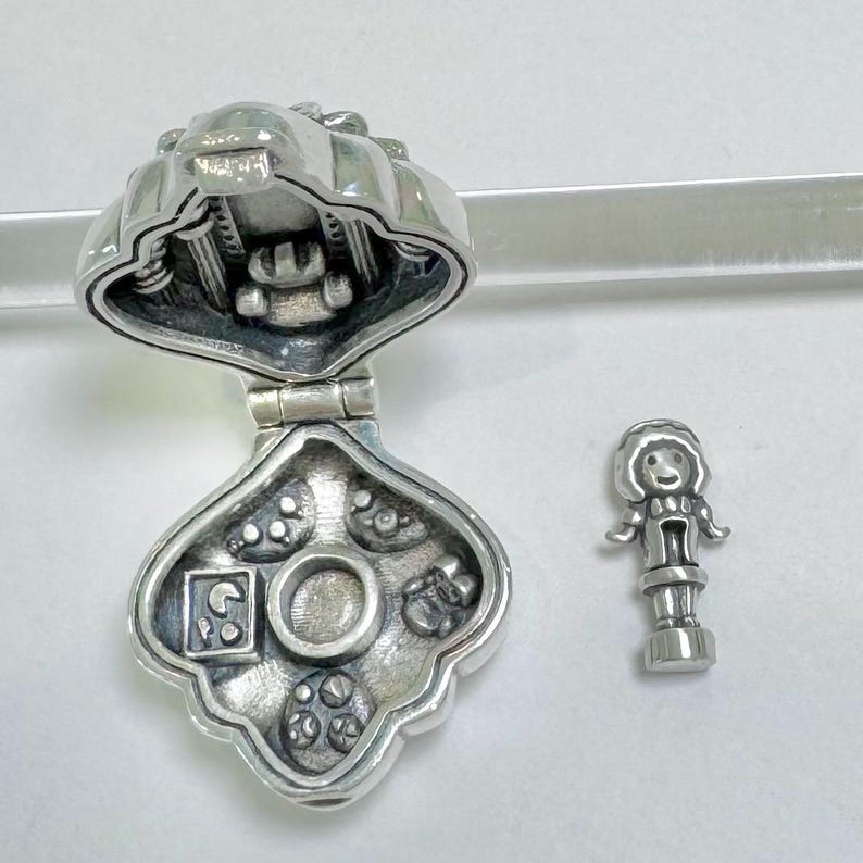 Pu&ograve; includere: Ciondolo in argento a forma di casa con un coperchio incernierato. L'interno della casa &egrave; decorato con un piccolo cerchio e quattro piccoli cerchi con disegni. Il ciondolo &egrave; aperto e un ciondolo in argento separato a forma di ragazza &egrave; accanto.