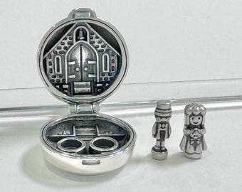 May include: A silver-toned locket with a church design on the lid, open to reveal two small ring holders. Two miniature figurines of a bride and groom stand beside the locket. The locket and figurines are highly detailed.