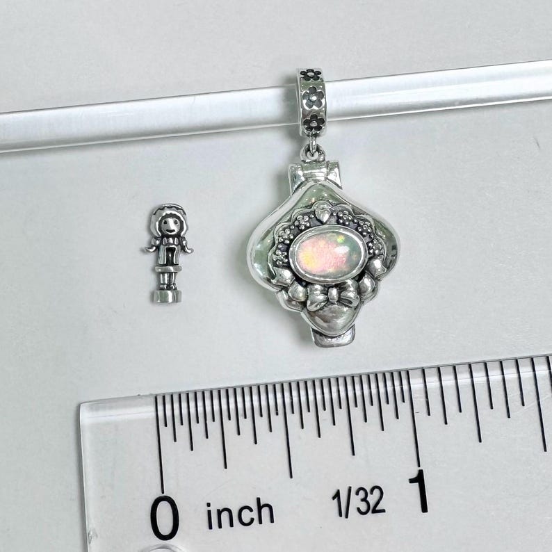 Pu&ograve; includere: Un ciondolo d'argento con un medaglione a forma di cuore che presenta una pietra di opale bianca e dettagli floreali. Il ciondolo &egrave; appeso a una catena d'argento e misura circa 1 pollice di lunghezza.