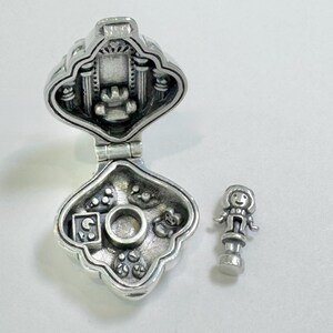 Pu&ograve; includere: Una scatola portaciondolo in argento con un coperchio incernierato, aperta per rivelare un piccolo interno con una bambola e altri ciondoli. La scatola ha la forma di un cuore e presenta un design dettagliato.