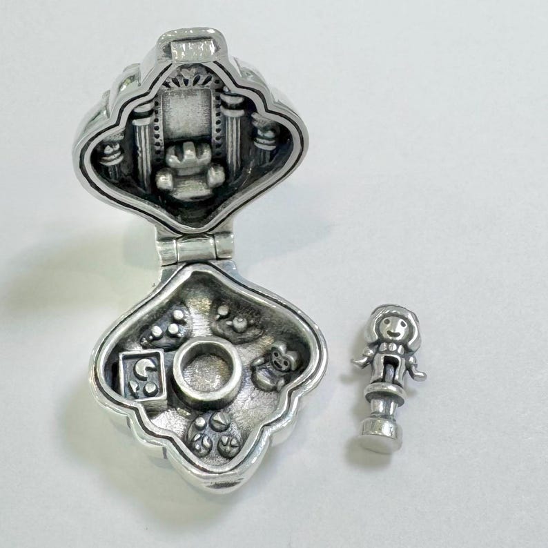 Pu&ograve; includere: Una scatola portaciondolo in argento con un coperchio incernierato, aperta per rivelare un piccolo interno con una bambola e altri ciondoli. La scatola ha la forma di un cuore e presenta un design dettagliato.